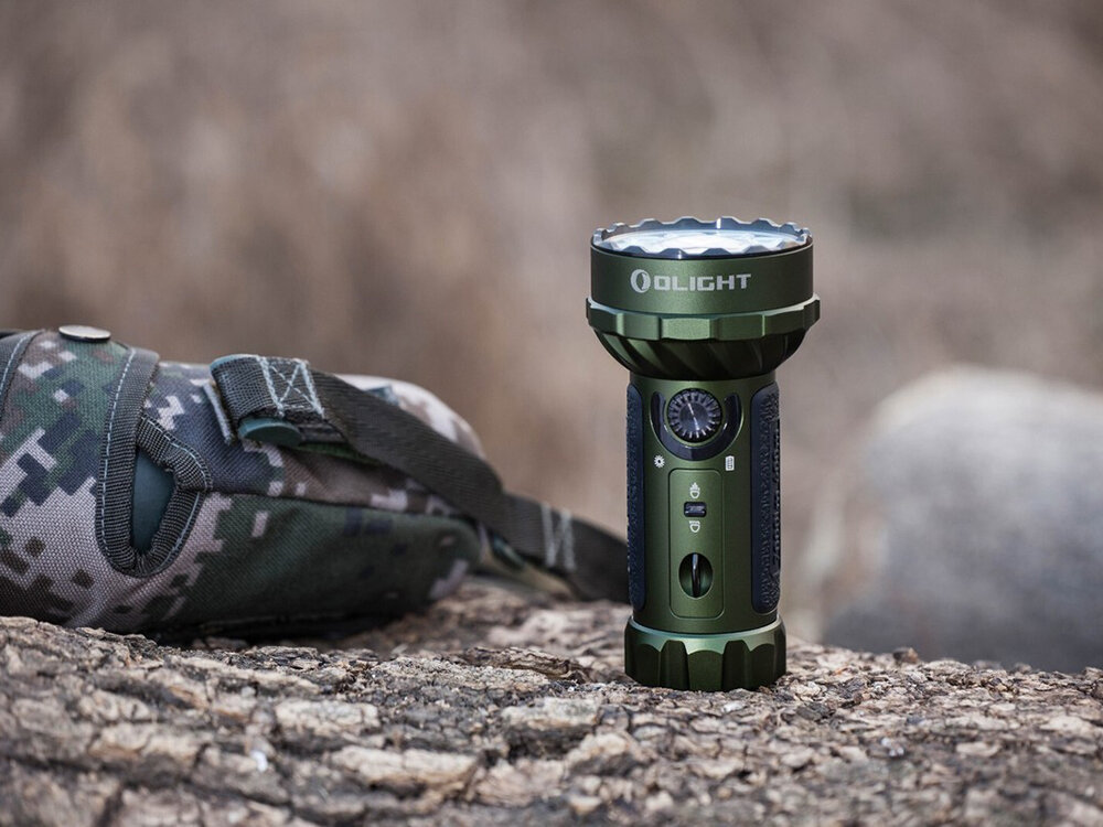 Latarka OLIGHT Marauder Mini QL000983 Zielony, 7000 lumenów, ergonomia, floodlight, RGB, Zielona latarka Olight Marauder Mini stoi pionowo na konarze, obok torba w kamuflażu, tło rozmyte.