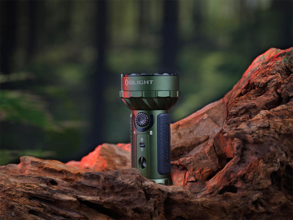 Latarka OLIGHT Marauder Mini QL000983 Zielony, aluminium, IPX8, odporna, O-ring, Olight Marauder Mini w zielonej wersji stoi na dużym kawałku kory, rozmyte tło leśne z mocnym światłem.