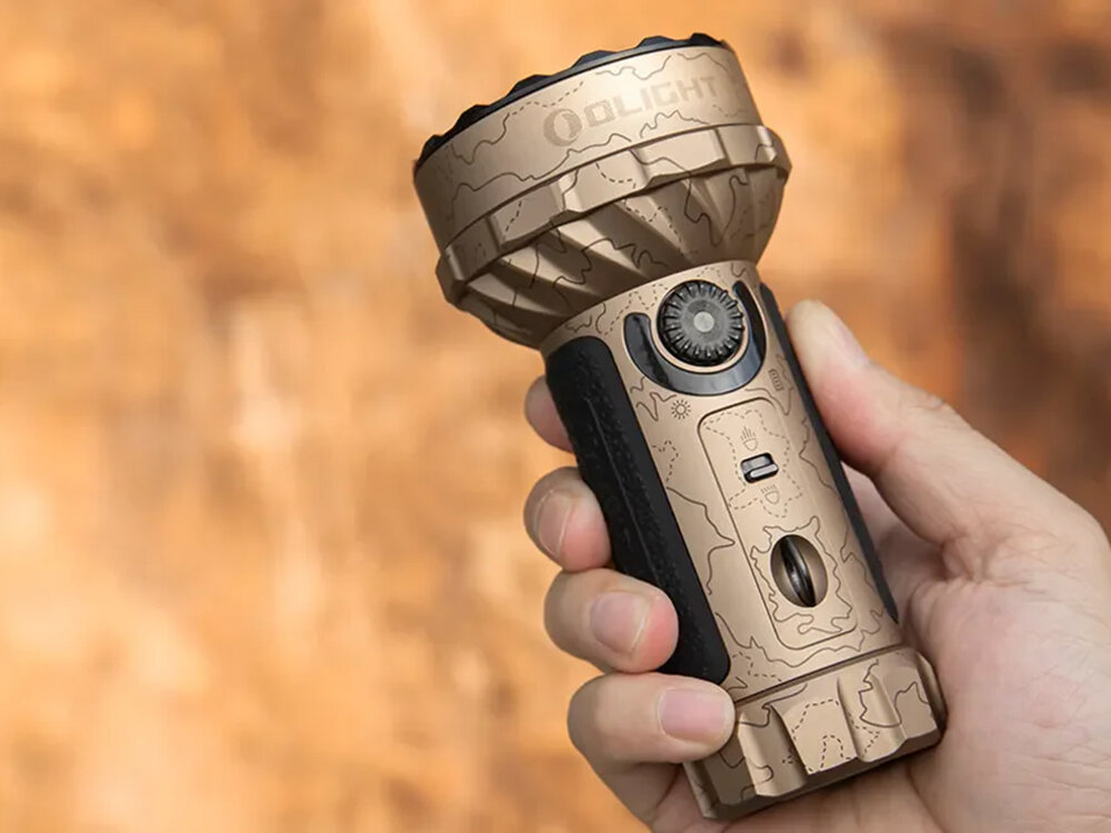 Latarka OLIGHT Marauder Mini QL691626 Piaskowy, aluminium, IPX8, odporna, O-ring, Olight Marauder Mini Desert w dłoni, wyraźna faktura, tło w odcieniach brązu, solidny uchwyt.