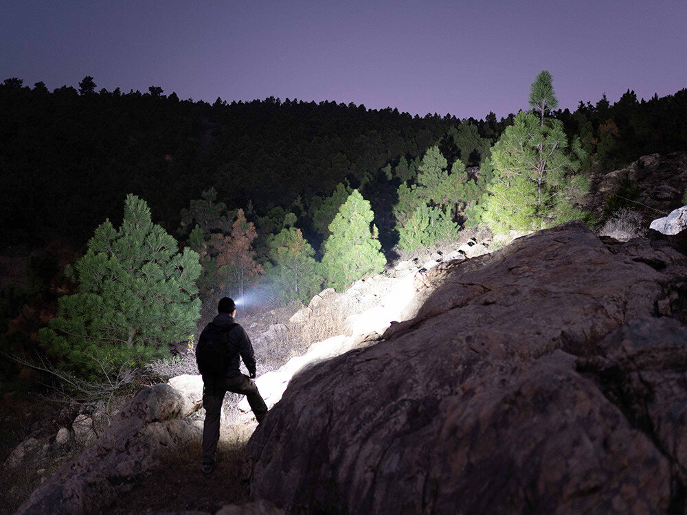 Latarka czołowa OLIGHT Perun 2 Mini QL000648, elektronika, moc, przegrzanie, ochrona, Nocna wyprawa w góry, użytkownik oświetla skarpę mocnym światłem latarki, zalesione tło.