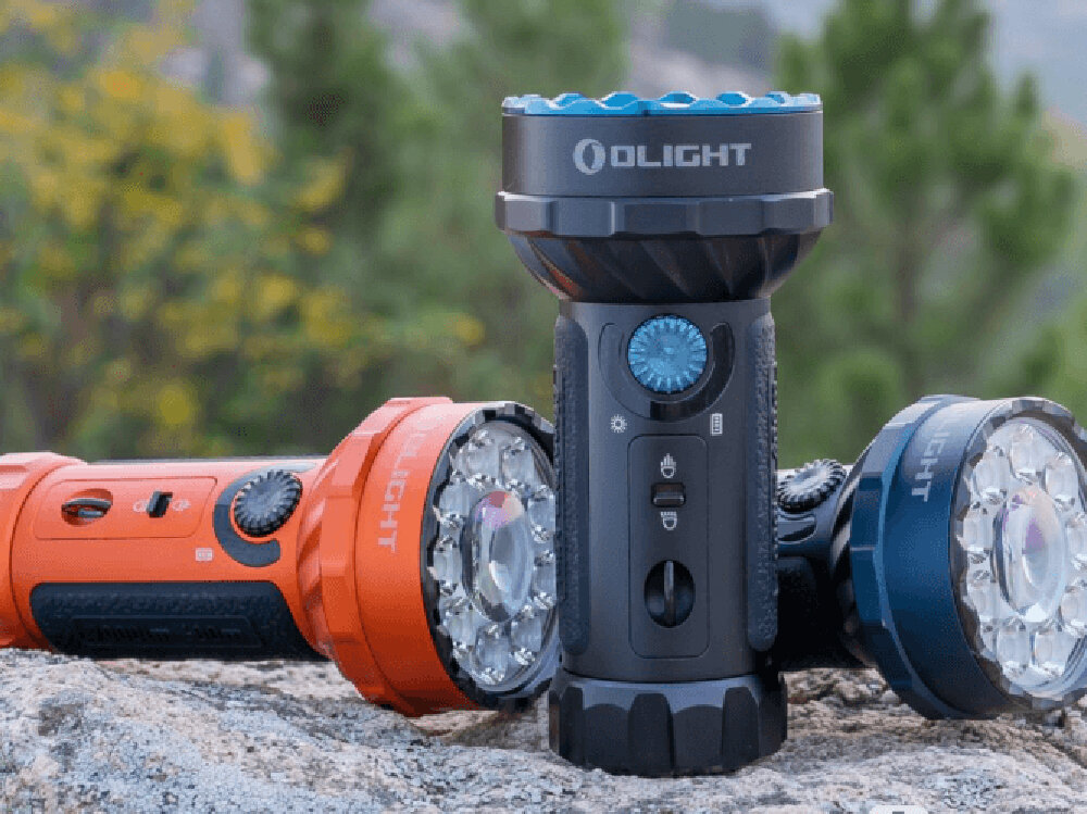 Latarka OLIGHT Marauder Mini QL000532 Pomarańczowy, 7000 lumenów, ergonomia, floodlight, RGB, Czarno-niebieska latarka Olight Marauder Mini stoi pionowo na kamieniu, obok leżą wersje pomarańczowa i niebieska, ostre tło z zielenią.