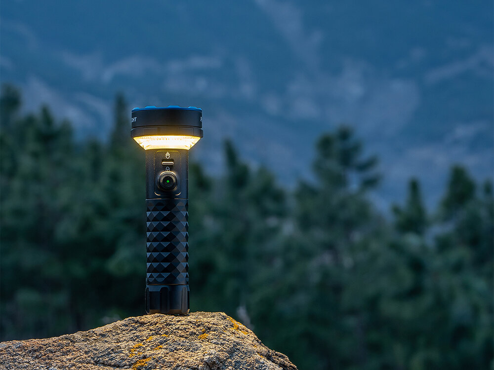 Latarka OLIGHT Prowess QL690681, Wszechstronne zastosowanie, tryby pracy, uchwyt, mocowania, Latarka Olight Prowess z ciepłym światłem, stoi na skale, góry i drzewa w tle.