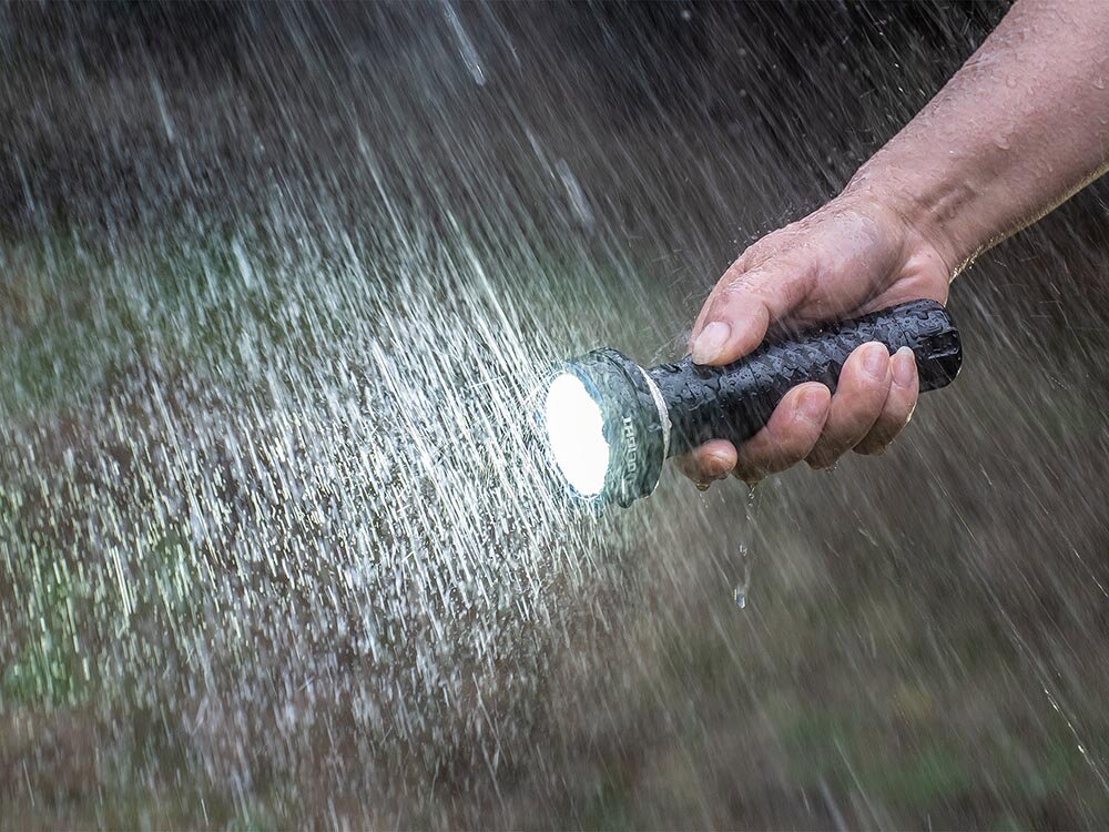 Latarka OLIGHT Prowess QL003670, Moc światła, zaufanie, profesjonalna latarka, warunki terenowe, Latarka używana w ulewnym deszczu, silne światło, dłoń trzyma korpus w strugach wody.
