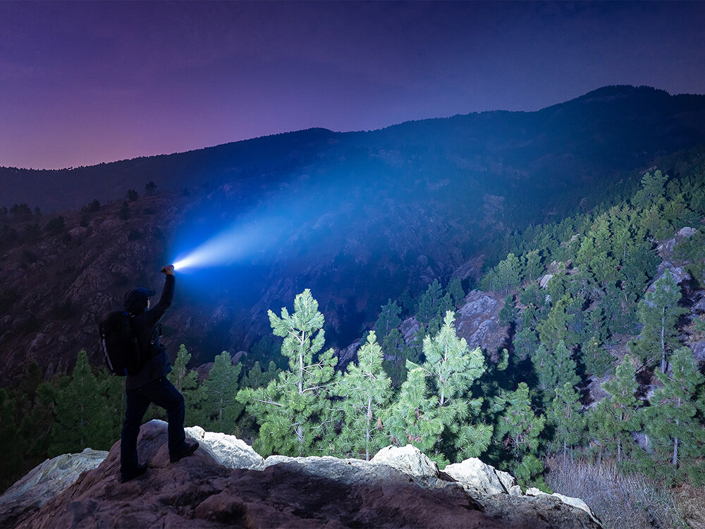 Latarka OLIGHT Prowess QL003670, Solidna konstrukcja, aluminium lotnicze, norma IPX7, odporność woda, Osoba na klifie nocą, mocna wiązka latarki Olight oświetla górski las w oddali.