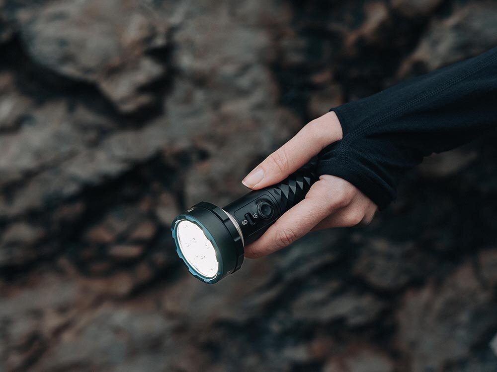 Latarka OLIGHT Prowess QL003670, Precyzyjna kontrola, przesuwany przycisk, regulacja jasności, szybkie przełączenie, Dłoń w czarnym rękawie trzyma włączoną latarkę Olight, tło skalne, detal uchwytu.