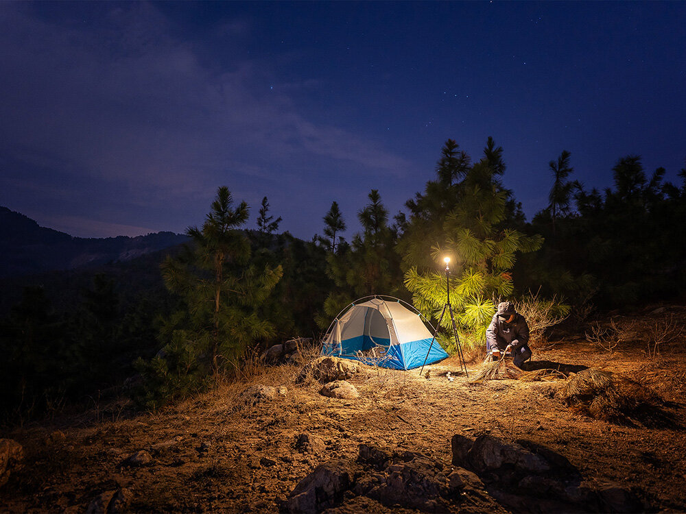 Latarka OLIGHT Prowess QL003670, Wskaźnik LED, montaż statyw, naklejki montażowe, elastyczne użytkowanie, Namiot oświetlony lampą na statywie, biwak nocą w lesie, osoba przygotowuje ognisko.