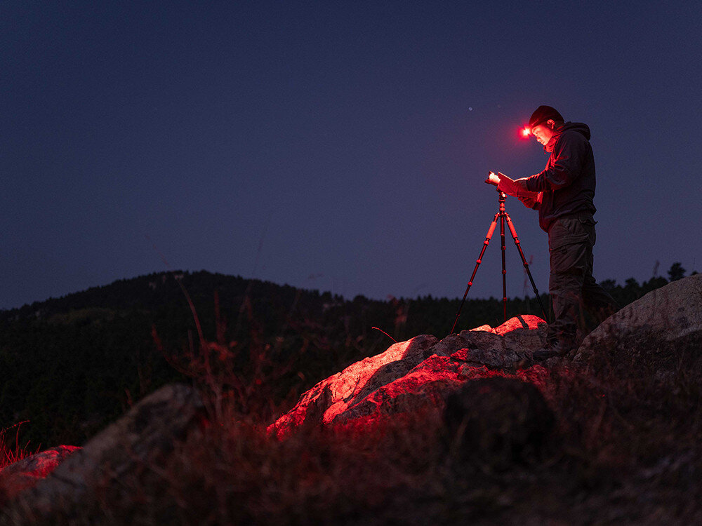 Latarka czołowa OLIGHT QL000594, tryby, czerwony, jasność, czas, Fotograf w terenie nocą, czerwone światło czołówki Olight, statyw na tle gór i nieba.