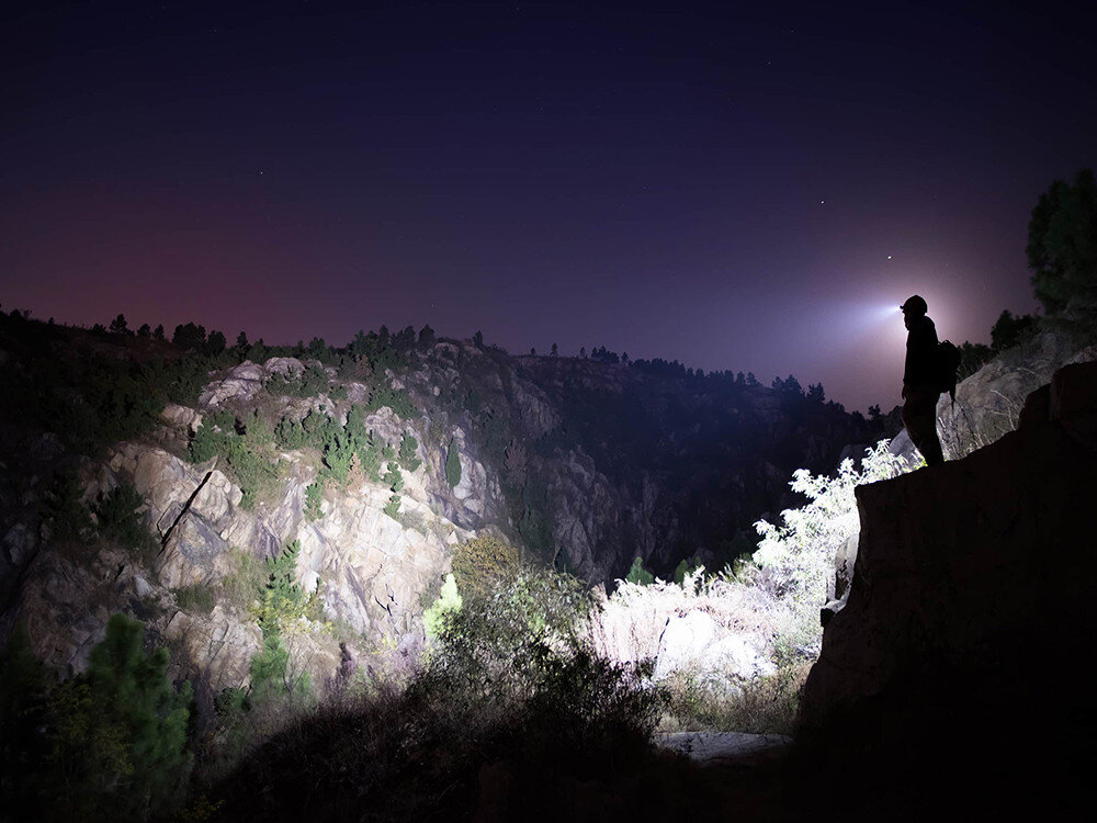 Latarka czołowa OLIGHT Perun 2 QL005063, opaska, montaż, kątowa, wygoda, Osoba z latarką na głowie oświetla kanion nocą, silny snop światła rozjaśnia skały i drzewa.