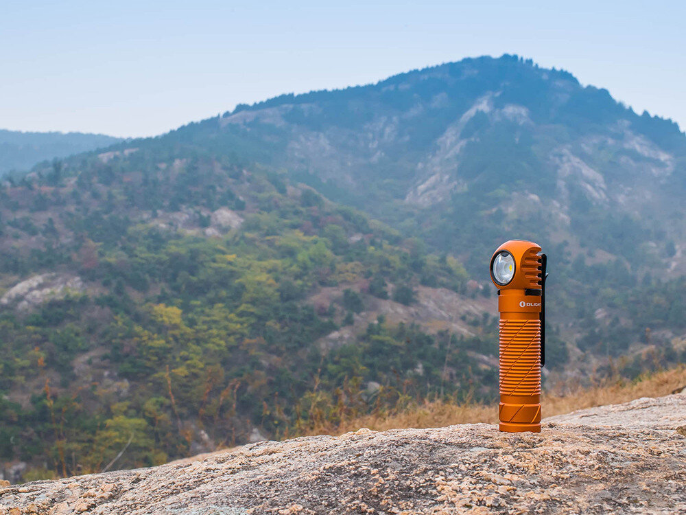 Latarka czołowa OLIGHT Perun 2 QL005063, magnes, klips, montaż, kieszeń, Pomarańczowa latarka Olight stojąca na tle gór i drzew, mocno wyeksponowana na pierwszym planie.
