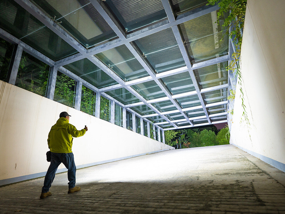 Latarka czołowa OLIGHT Perun 3 QL690193, aluminium, anodyzacja, IP68, wytrzymałość, Mężczyzna w jasnej kurtce oświetla szeroki tunel latarką, noc, nowoczesna architektura.