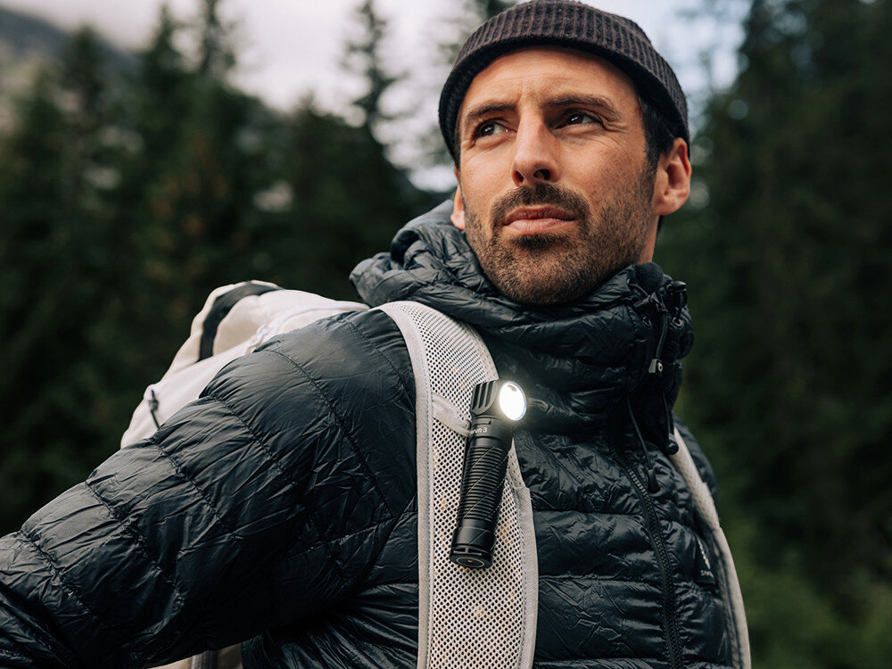 Latarka czołowa OLIGHT Perun 3 QL690193, opaska, klips, magnes, montaż, Mężczyzna podczas trekkingu z latarką przypiętą do ramienia plecaka, górski las w tle.