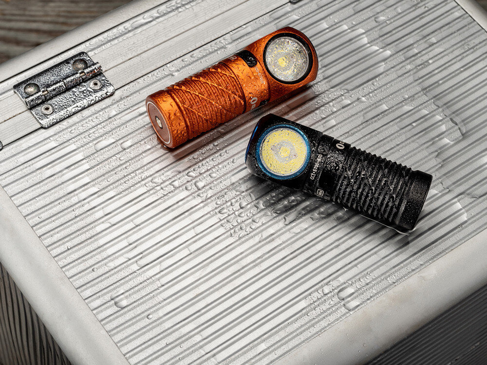 Latarka czołowa OLIGHT Perun 3 Mini CW QL690759, system, elektroniczny, zabezpieczenia, przycisk, Pomarańczowa i czarna latarka Olight pokryte wodą, leżą na aluminiowej skrzynce, odporność IPX8.