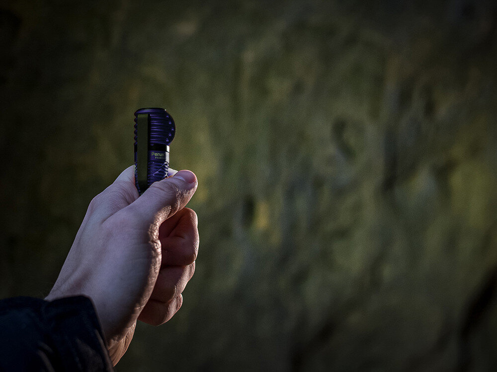 Latarka czołowa OLIGHT Perun 3 Mini QL692838, system, zabezpieczenia, diody, przycisk, Ręka trzyma pionowo fioletową latarkę Olight Perun 3 Mini, skała w tle, ujęcie w cieniu.