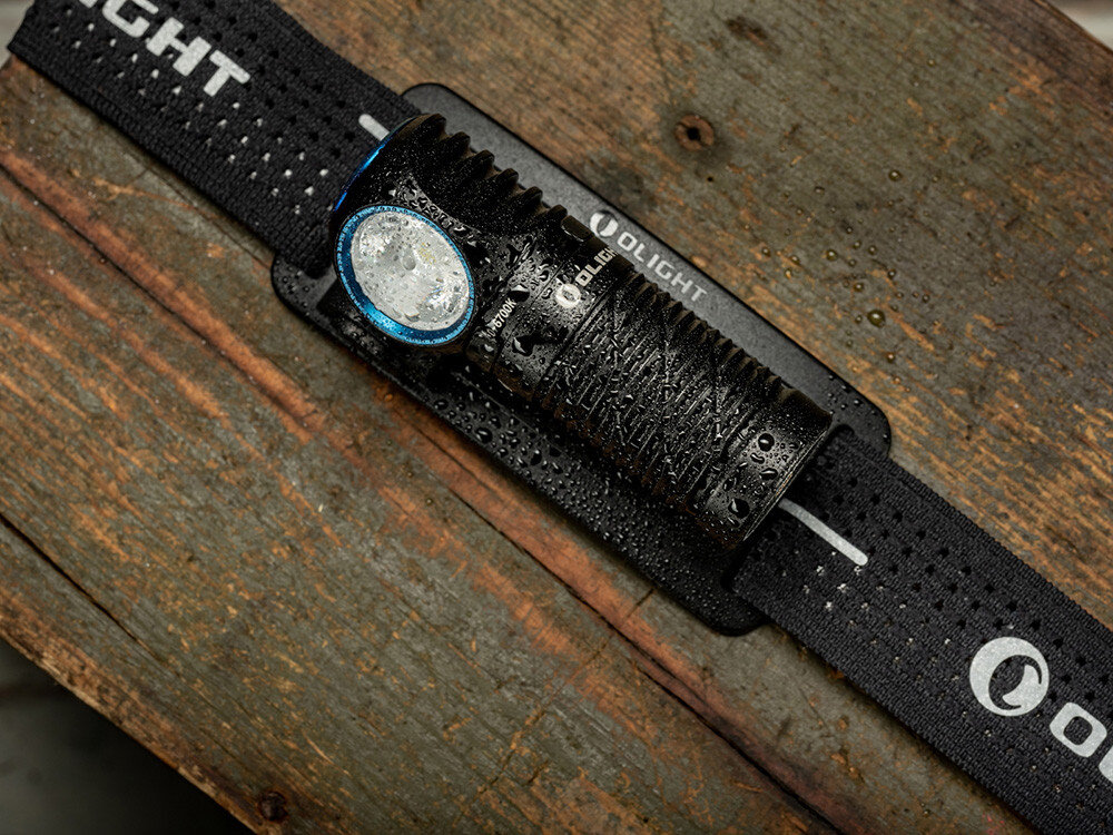 Latarka czołowa OLIGHT Perun 3 Mini QL692401, aluminium, anodyzacja, IPX8, odporność, Czarna latarka Olight z opaską, pokryta kroplami wody, leży na drewnianej desce, wytrzymałość.