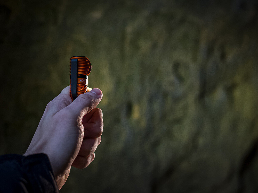 Latarka czołowa OLIGHT Perun 3 Mini QL692418, system, elektroniczny, zabezpieczenia, przycisk, Ręka trzyma pomarańczową latarkę Olight Perun 3 Mini, rozproszony światłocień, tło skała.