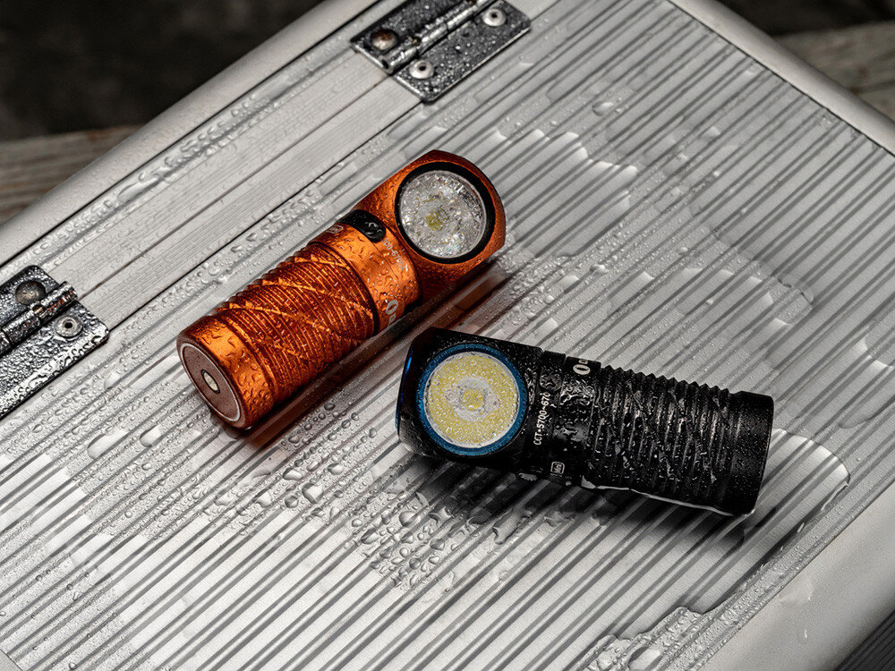 Latarka czołowa OLIGHT Perun 3 Mini QL692418, akumulator, magnetyczna, ładowarka, wskaźnik, Dwie latarki Olight (czarna i pomarańczowa) pokryte kroplami wody na skrzynce aluminiowej.