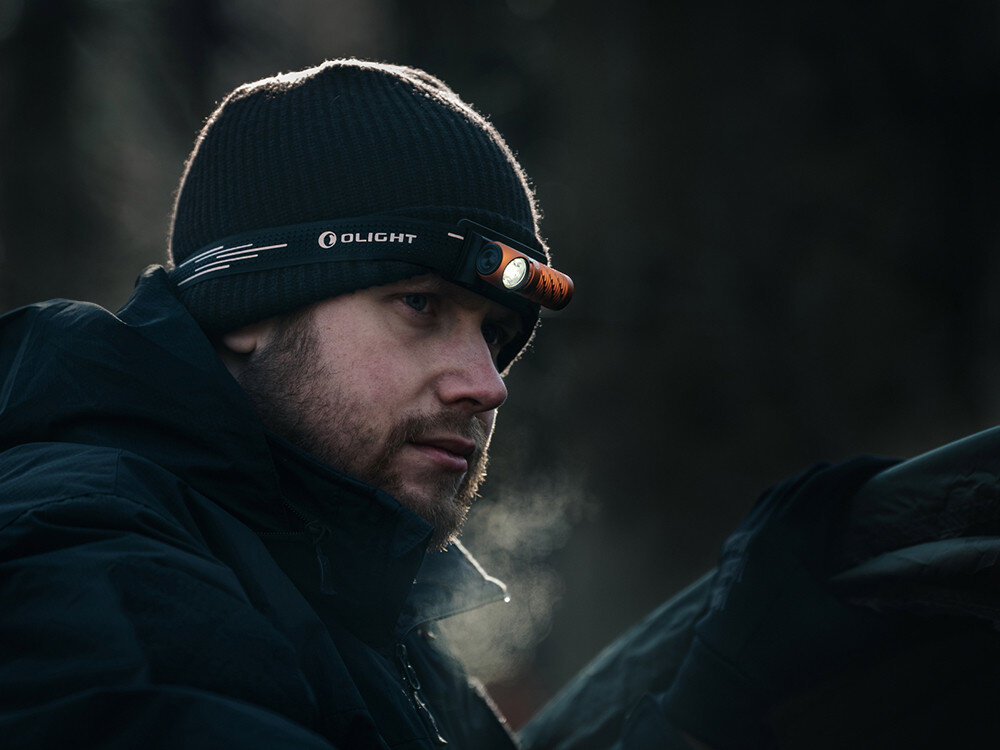 Latarka czołowa OLIGHT Perun 3 Mini QL692418, opaska, regulacja, odblask, komfort, Mężczyzna w czapce Olight z pomarańczową czołówką patrzy w bok, leśne tło, zimny poranek.