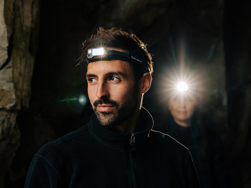 Latarka czołowa OLIGHT Array 2 Pro QL128782, tryby, Flood, Spot, wiązka, Mężczyzna w jaskini, latarka czołowa Olight Array 2 Pro świeci mocnym światłem, ciemne otoczenie.