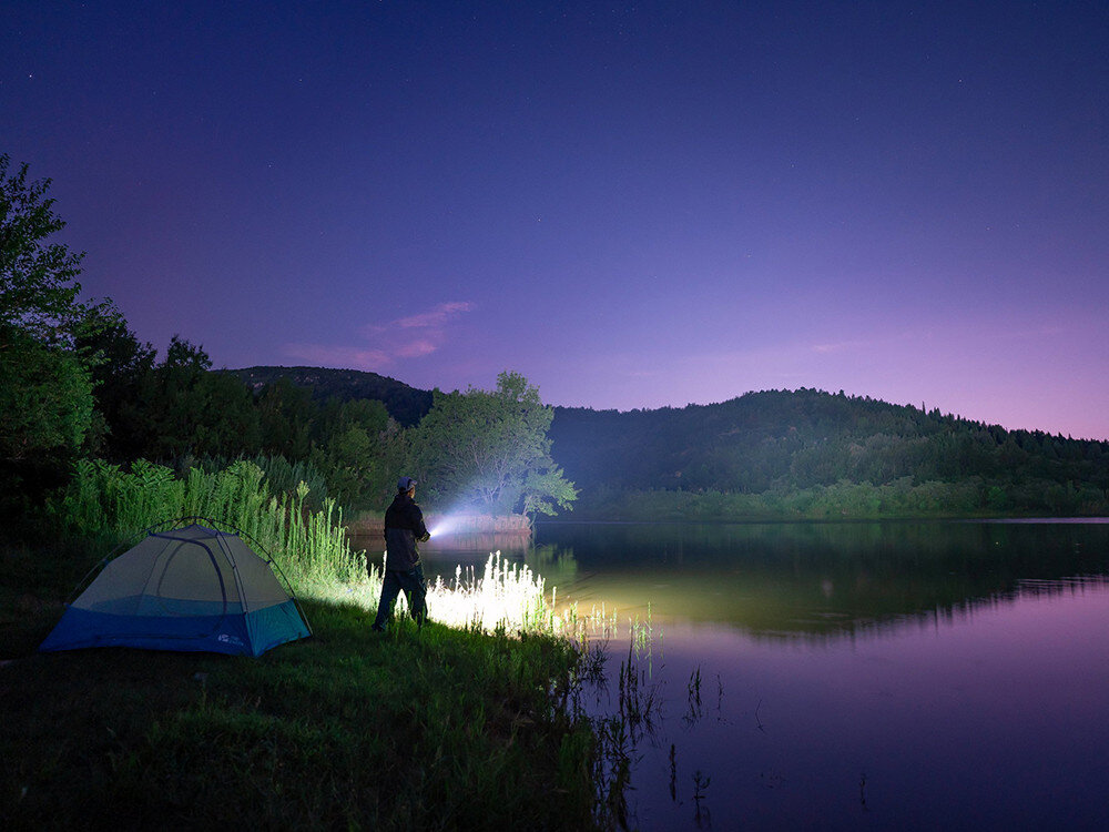 Latarka OLIGHT Seeker 4 Pro QL006169, akumulator, ładowanie, usb, zabezpieczenie, Camper nad jeziorem po zmroku, latarka Olight oświetla brzeg i wodę, namiot w rogu kadru.