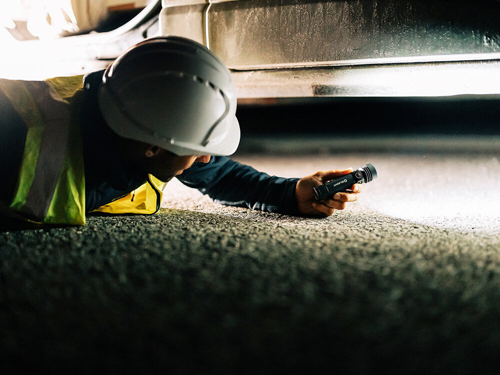 Latarka OLIGHT Seeker 4 Pro QL006152, tryby, turbo, jasność, czas, Pracownik w kasku, leżąc pod pojazdem, oświetla asfalt mocną latarką Olight, inspekcja nocą.