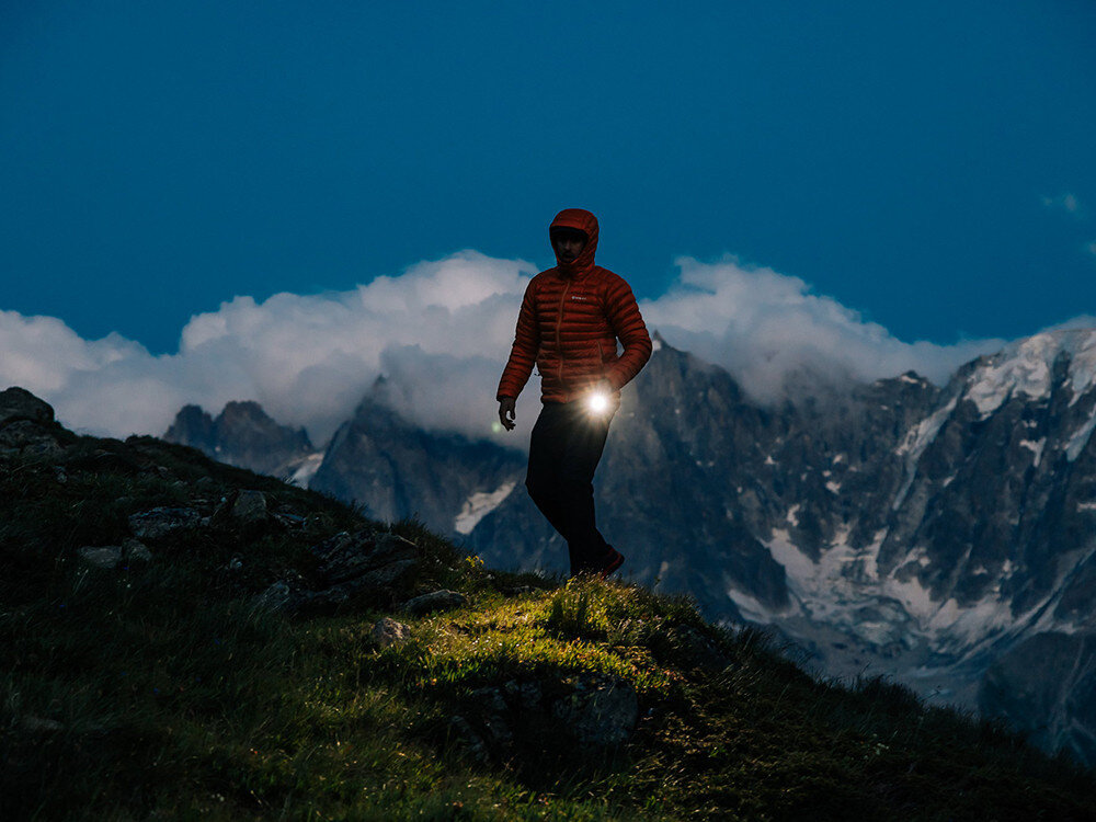 Latarka OLIGHT Seeker 4 Pro QL692197, sterowanie, przycisk, energia, diody, Osoba w ciepłej kurtce na tle gór i chmur, latarka Olight oświetla trawę przy zejściu zboczem.