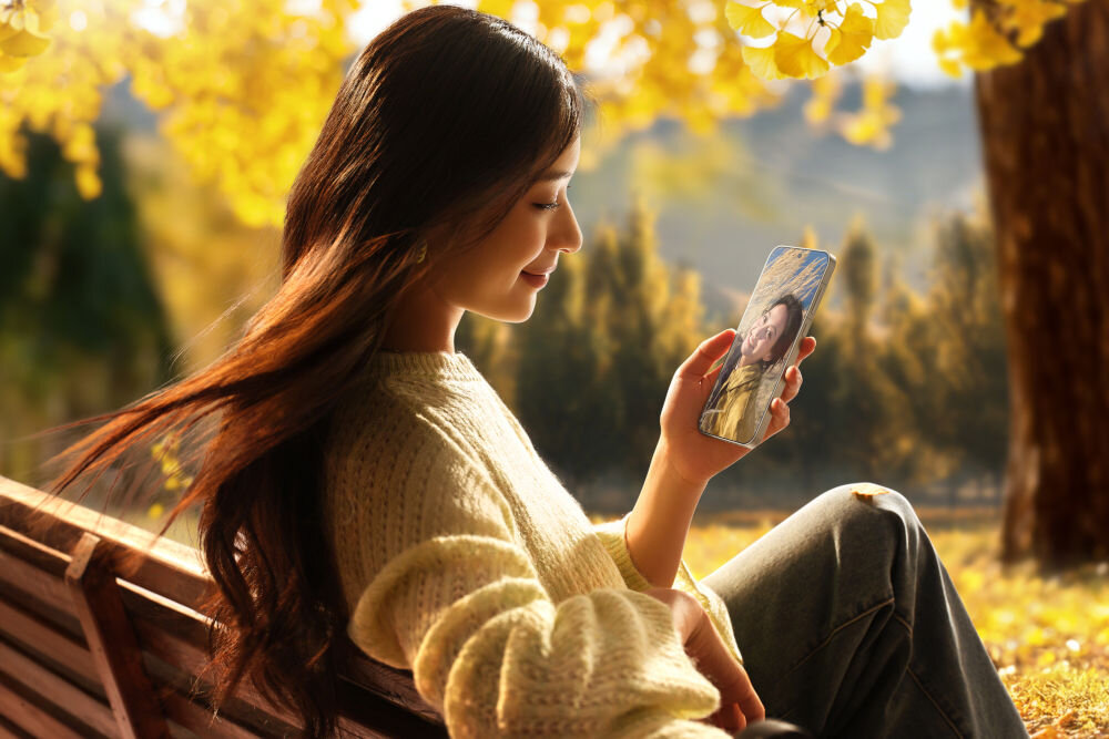 OPPO Find X9 Pro wyświetlacz 6,78 cala AMOLED ProXDR 120 Hz 3600 nitów Gorilla Glass Victus 2 Młoda kobieta z długimi, brązowymi włosami, siedząca na ławce w jesiennym parku, ubrana w jasny, wełniany sweter. Uśmiecha się, trzymając smartfon OPPO, na którego ekranie widać zbliżenie twarzy mężczyzny. W tle widać jaskrawożółte liście i drzewa.