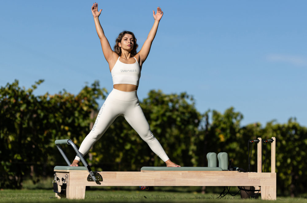Reformer do pilatesu TOORX Liveness PTX3000 ogród osoba ćwiczy na reformerze stoi na wózku niebo drzewa wózek o wymiarach 600 × 1025 mm osiem kółek z poliuretanu łożyska wałeczkowe płynne przemieszczanie przesuw wynoszący około 820 mm prowadnice ze stali nierdzewnej