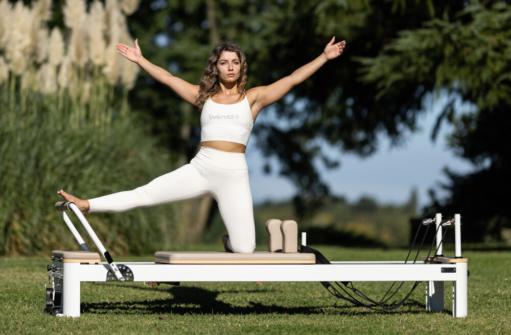 Osoba ćwiczy na reformerze do pilatesu na zewnątrz, ubrana w jasny strój sportowy z napisem \'Liveness\'. W tle widać drzewa i trawę.
