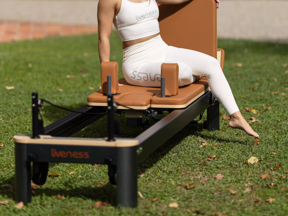 Reformer do pilatesu TOORX Liveness PTX7500 Brązowo-czarny ogród trawnik osoba siedzi na reformerze z aluminium w kolorze Underwood&Earth wysoka odporność na intensywną eksploatację do 150 kg zgodność z normami EN ISO 20957-1 oraz EN ISO 20957-2 w klasie S