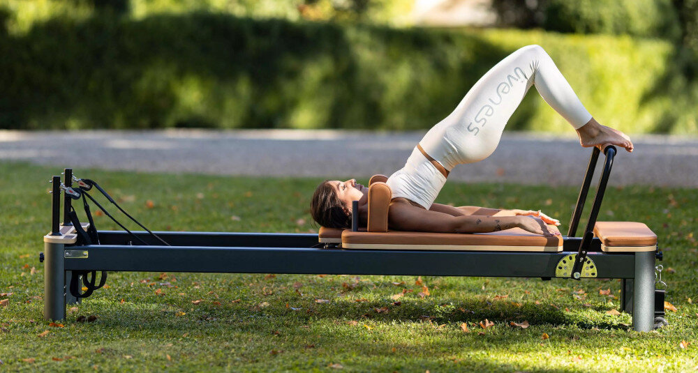 Reformer do pilatesu TOORX Liveness PTX7500 Brązowo-czarny ogród trawnik osoba ćwiczy na reformerze stojąc wózek o wymiarach 600 × 944 mm osiem poliuretanowych kółek łożyska przesuw do 1055 mm ruch pozostaje płynny i stabilny wysokość około 412 mm od podłogi