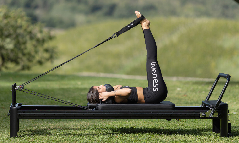 Reformer do pilatesu TOORX Liveness PTX7500 Czarny ogród trawnik osoba ćwiczy na reformerze stojąc wózek o wymiarach 600 × 944 mm osiem poliuretanowych kółek łożyska przesuw do 1055 mm ruch pozostaje płynny i stabilny wysokość około 412 mm od podłogi