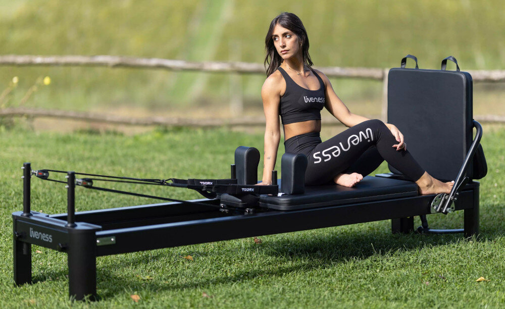 Reformer do pilatesu TOORX Liveness PTX7500 Czarny ogród trawnik osoba siedzi na reformerze regulowane paski z podwójnymi pętlami uchwyty pierścienie pasek na stopy box jumpboard do treningów wzmacniających rozciągających ćwiczeń rozwijających koordynację i dynamikę ruchu