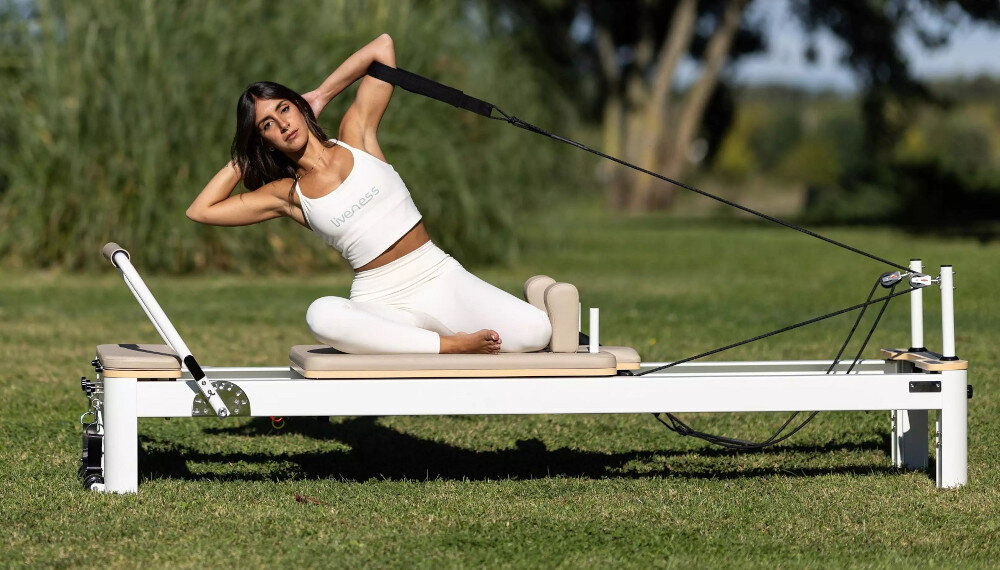 Reformer do pilatesu TOORX Liveness PTX7500 Brązowy ogród trawnik osoba ćwiczy na reformerze stojąc wózek o wymiarach 600 × 944 mm osiem poliuretanowych kółek łożyska przesuw do 1055 mm ruch pozostaje płynny i stabilny wysokość około 412 mm od podłogi