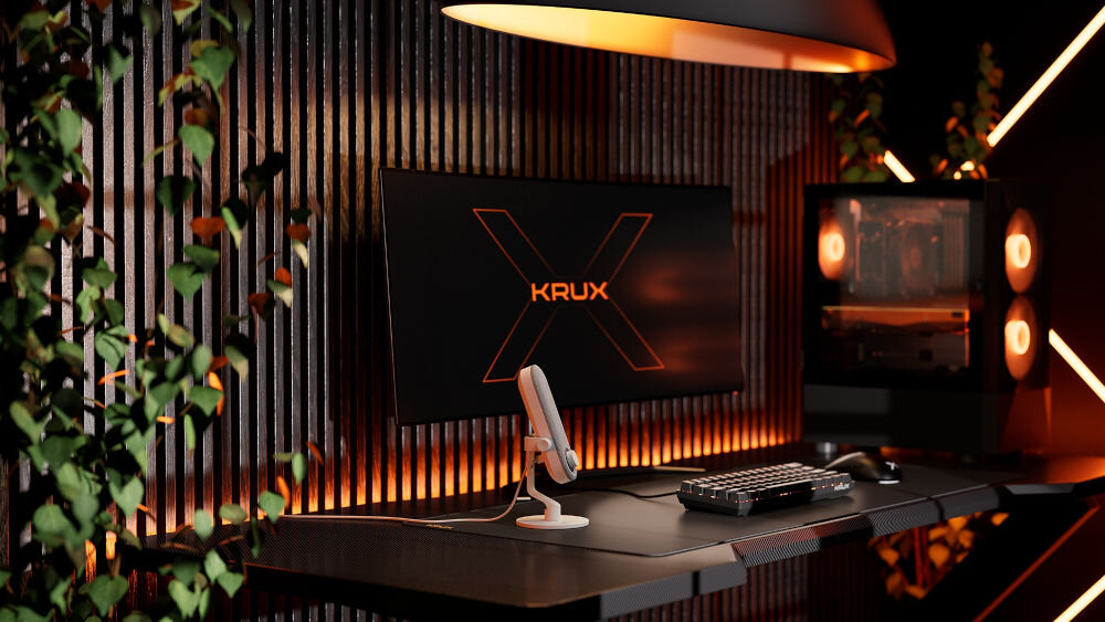 Mikrofon KRUX Pin KRXC019 Kardioidalny Biały Pełne ujęcie stanowiska gamingowego z mikrofonem KRUX Pin na pierwszym planie, monitor z logo KRUX w tle, oświetlenie RGB w odcieniach pomarańczowo-czerwonych oraz bujna roślina po lewej stronie