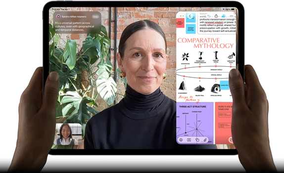 Widoczny od przodu ustawiony poziomo iPad Pro w kolorze gwiezdnej czerni, zaokrąglone rogi, czarna ramka wyświetlacza. Ręce trzymają urządzenie z lewej i prawej strony, na ekranie tłumaczona na żywo rozmowa przez FaceTime. Widać też aplikację edukacyjną z prezentacją „Comparative Mythology”, z tekstem, diagramami i odręcznymi notatkami