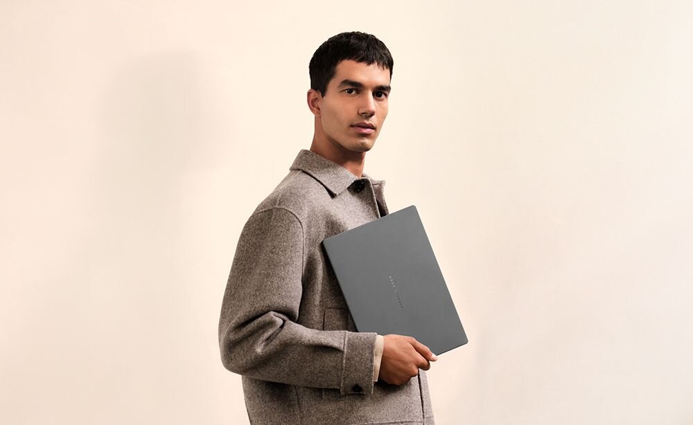 Mężczyzna w szarym swetrze trzymający laptop ASUS Zenbook w kolorze szarym na minimalistycznym beżowym tle, demonstracja eleganckiego designu i smukłej konstrukcji laptopa premium - Design funkcjonalny w podróży