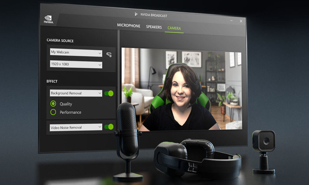 Na ekranie komputera widoczny jest interfejs programu NVIDIA Broadcast z zakładką \'CAMERA\', gdzie widać podgląd z kamery internetowej. Przed monitorem znajdują się mikrofon, słuchawki i mała kamera.