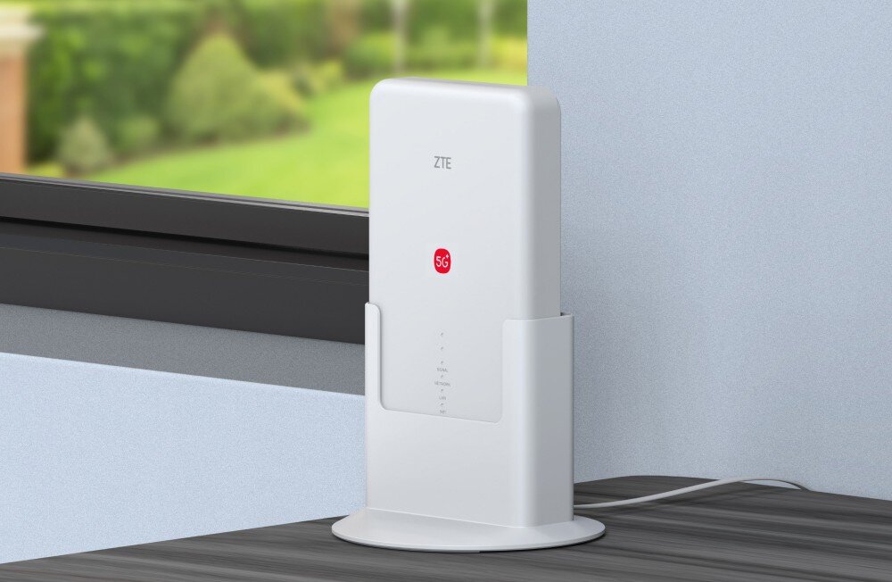 Router ZTE G5 Max Pro Biała wewnętrzna jednostka (IDU) routera ZTE 5G stoi pionowo w podstawce na drewnianym biurku/parapecie, przy ścianie i oknie, za którym widać rozmyty ogród. Urządzenie z logo ZTE i 5G prezentuje dyskretny i elegancki design (5G CPE), idealnie wpasowujący się w dom, aby zapewnić szybki bezprzewodowy Internet wewnątrz pomieszczenia.