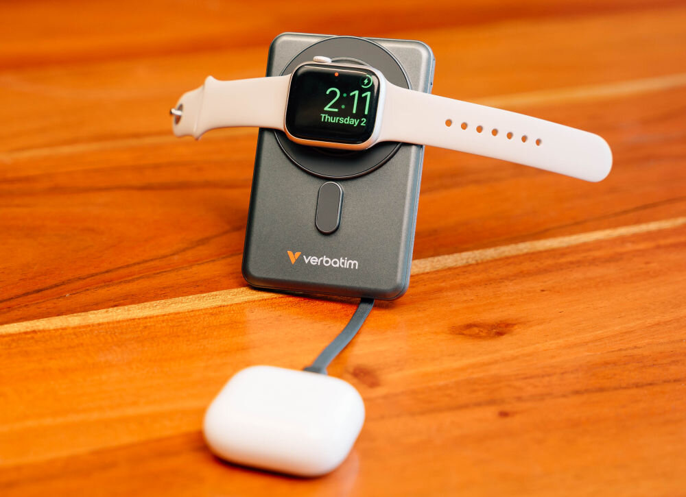 Na drewnianej powierzchni znajduje się szary powerbank z logo \'verbatim\', na którym umieszczony jest smartwatch z wyświetlaczem pokazującym godzinę 2:11 i datę \'Thursday 2\'. Obok powerbanka leży biały element z kablem.