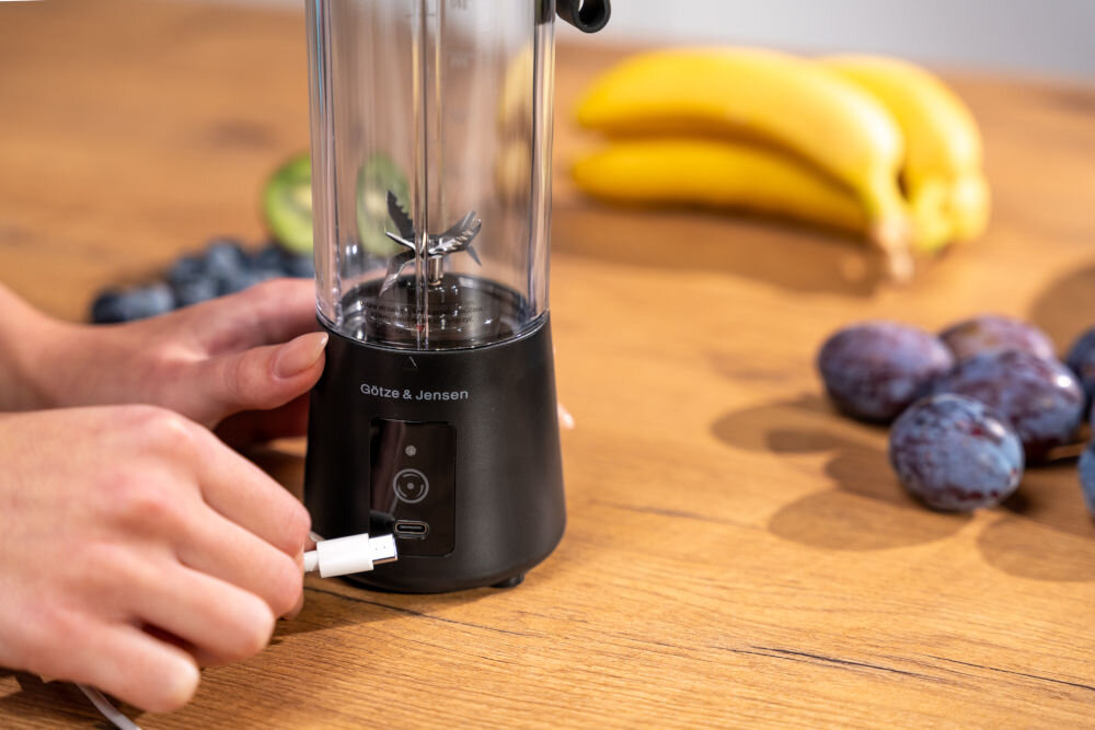 Osoba podłącza kabel do czarnego blendera personalnego marki Götze & Jensen, który stoi na drewnianym blacie. W tle widoczne są owoce, takie jak banany i śliwki.