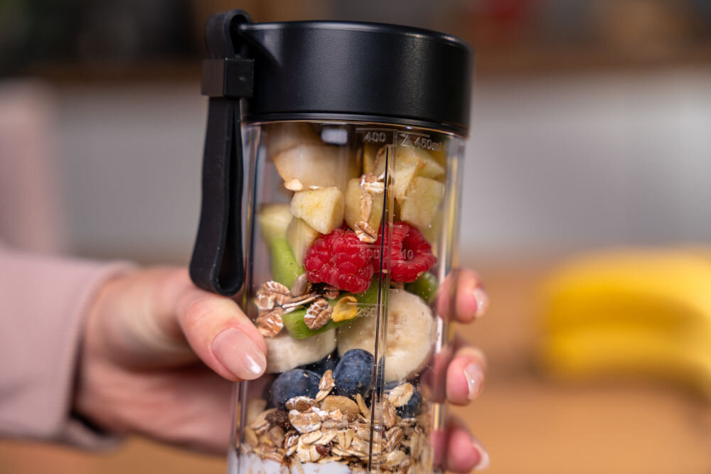 Osoba trzyma przezroczysty pojemnik blendera wypełniony owocami, płatkami i jogurtem. Na pojemniku widoczne są miarki z oznaczeniami 450 ml i 16 oz.