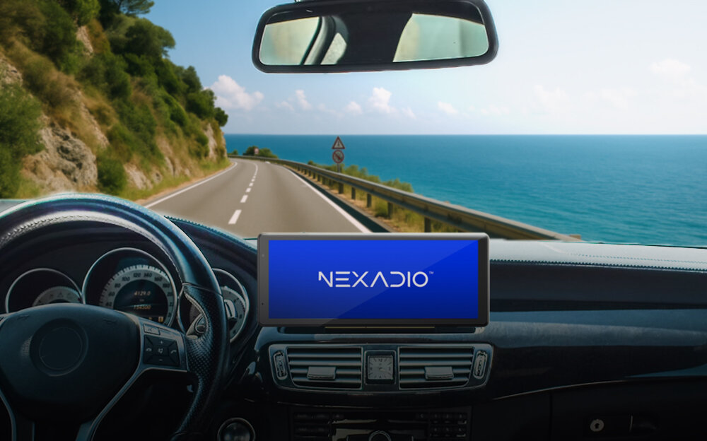 Wnętrze samochodu z widocznym kierownicą i deską rozdzielczą, na której znajduje się tablet z logo \'NEXADIO\'. Za szybą widać drogę biegnącą wzdłuż wybrzeża z widokiem na morze.