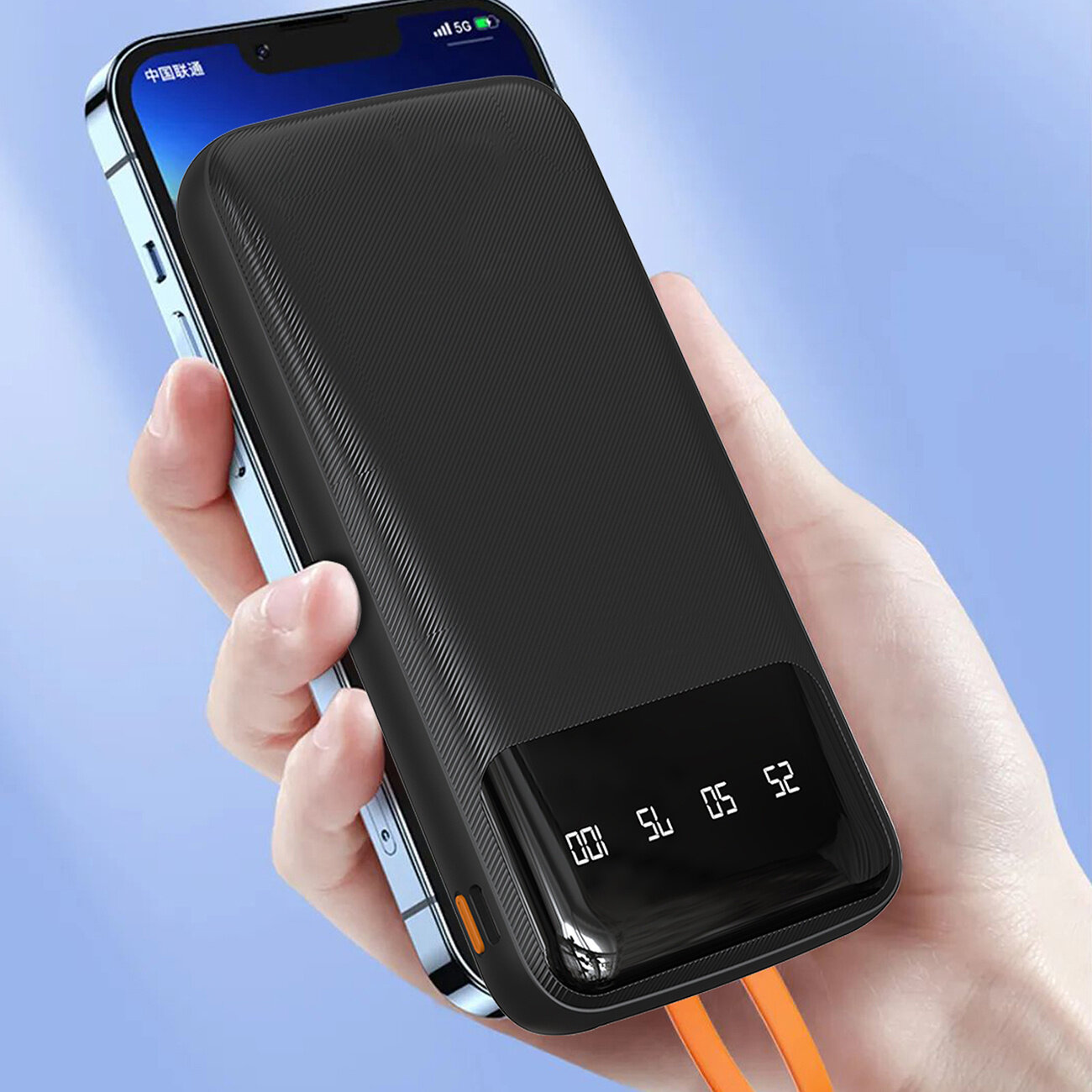 Osoba trzyma czarny powerbank z wyświetlaczem pokazującym cyfry 00, 54, 05, 52. Powerbank ma wbudowane kable w kolorze pomarańczowym.