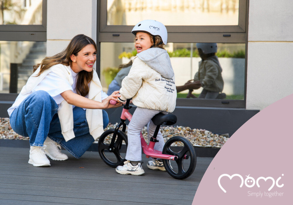 Rowerek biegowy MOMI Isoka Różowy ogród taras dziecko siedzi na rowerku obok dorosła osoba klęczy przed rowerkiem w tle okno włączone światła na kółkach aktywność rozwój pierwszy pojazd dziecka