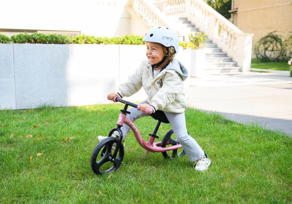 Rowerek biegowy MOMI Isoka Różowy ogród trawnik dziecko siedzi na rowerku w tle biały dom schody do domu lekka rama o masie 3,2 kg swobodne prowadzenie stalowa 9,5-calowa konstrukcja trwałość stabilność ramy wspiera rozwój równowagi