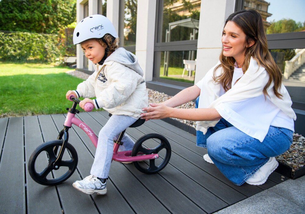 Rowerek biegowy MOMI Isoka Różowy ogród trawnik dziecko siedzi na rowerku za dzieckiem klęczy dorosła osoba trzyma dłonie na siodełku w tle okna domu miękkie regulowane siedzisko o zakresie 30–43 cm można dopasować wysokość do wzrostu dziecka prosta regulacja prawidłowa postawa lepsze panowanie nad rowerkiem