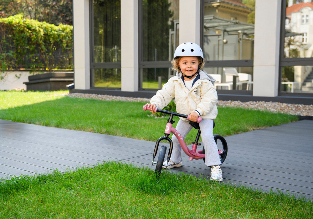 Rowerek biegowy MOMI Isoka Różowy ogród trawnik dziecko siedzi na rowerku w tle okna domu drzewa szeroka kierownica z gumowymi, antypoślizgowymi uchwytami regulacja wysokości kierownicy od 49 do 54 cm bez użycia narzędzi rowerek rośnie razem z dzieckiem
