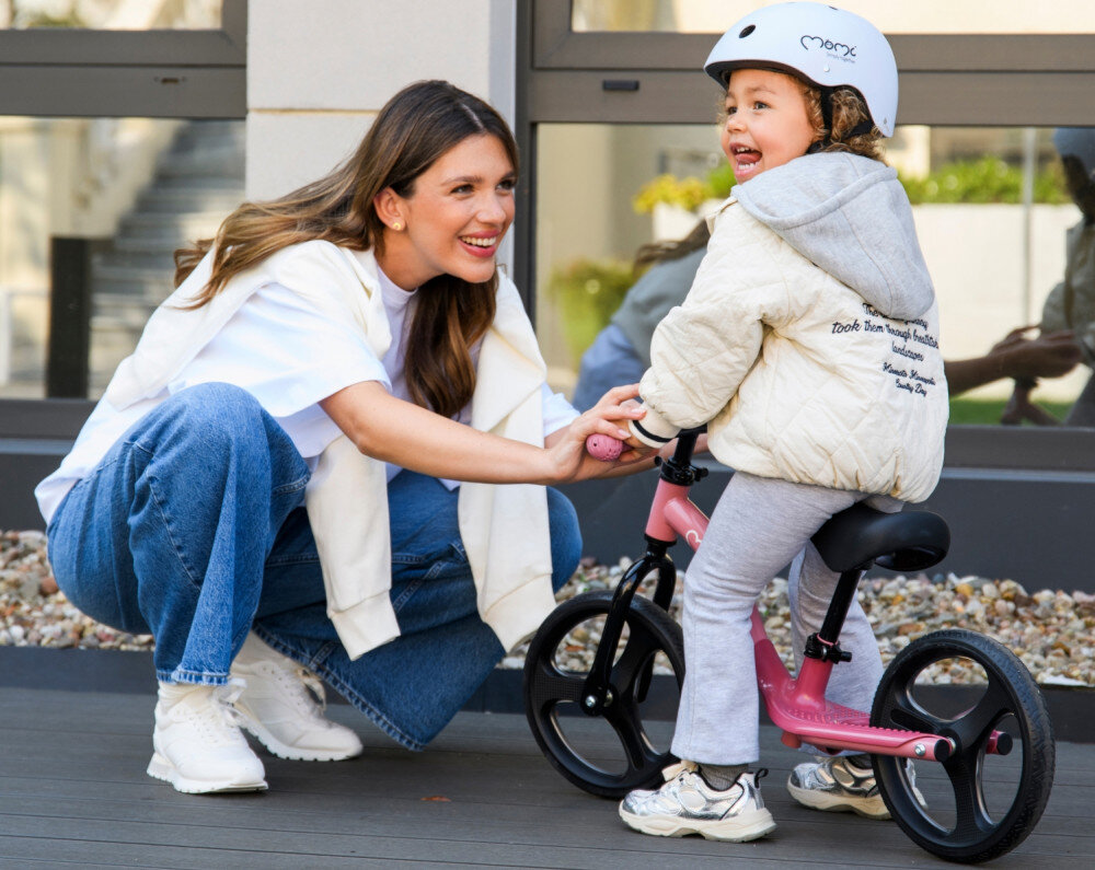 Rowerek biegowy MOMI Isoka Różowy 10-calowe ogród taras dziecko siedzi na rowerku obok dorosła osoba klęczy przed rowerkiem w tle okno koła wykonane z termoplastycznej gumy TPR trwałość elastyczność skutecznie niwelują wstrząsy płynna i wygodna jazda odporność na ścieranie bezpiecznie i komfortowo można rozwijać równowagę oraz koordynację ruchową