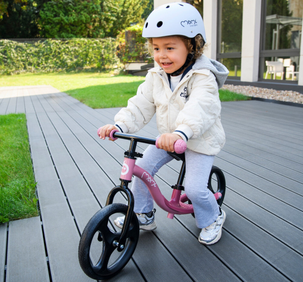 Rowerek biegowy MOMI Isoka Różowy taras ogród trawnik dziecko siedzi na rowerku spełnia europejskie normy EN 71-1, EN 71-2 i EN 71-3 bezpieczeństwo zastosowanych materiałów i konstrukcji odpowiedni dla dzieci od 2. roku życia do maksymalnej wagi do 35 kg sprawdzony pod kątem użytkowania