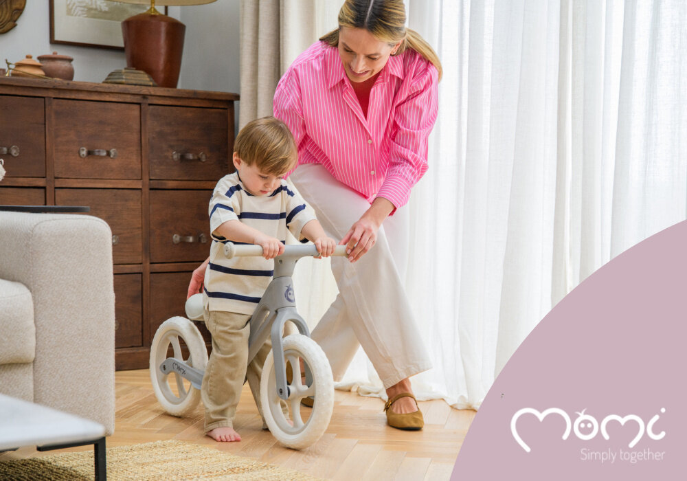 Rowerek biegowy MOMI Noru Szary pokój dziecko siedzi na rowerku obok dorosła osoba trzyma kierownicę rowerka w tle okno komoda meble aktywność rozwój pierwszy pojazd dziecka